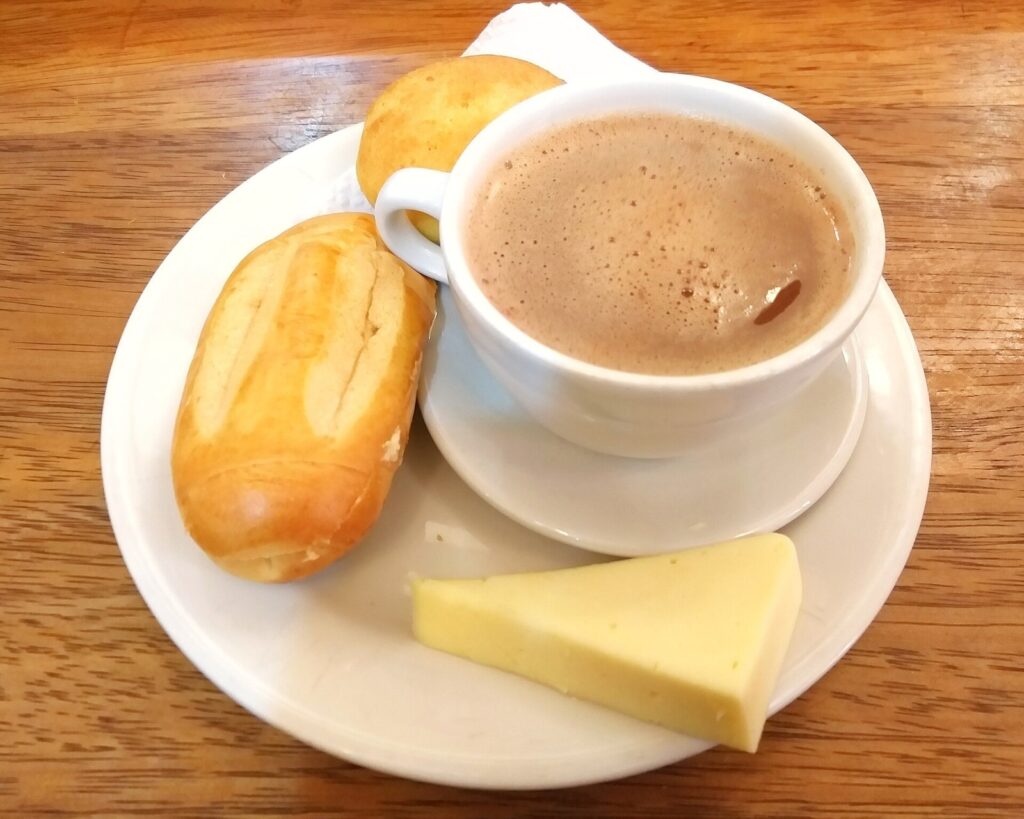Taza de chocolate caliente acompañada de pan, almojábana y una tajada de queso sobre un plato blanco