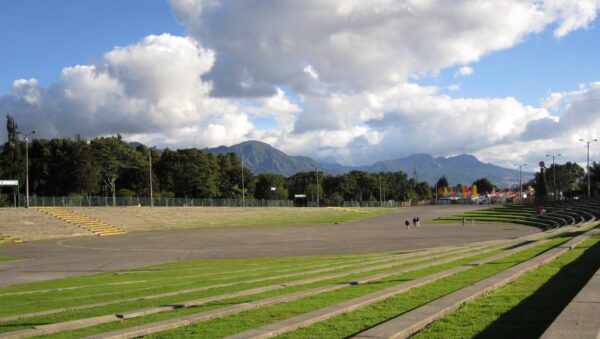Gradería verde y explanada del escenario principal del Parque Simón Bolívar en Bogotá bajo un cielo azul con nubes, con los cerros orientales al fondo