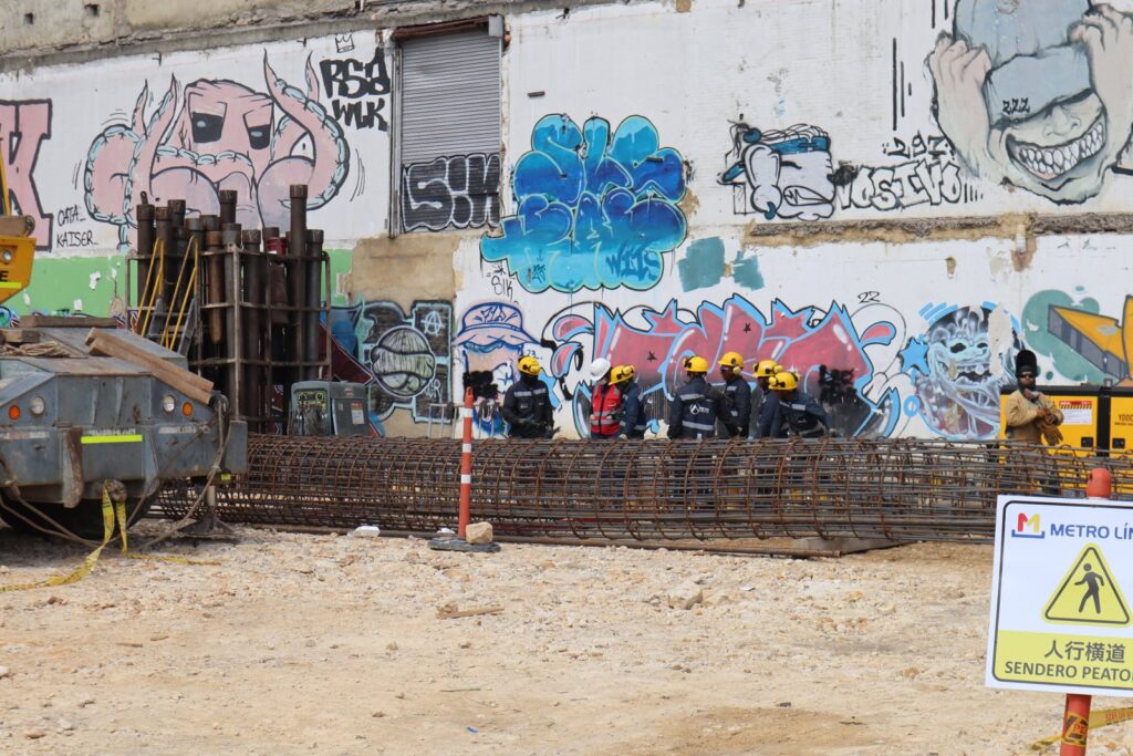 Obras de la estación Avenida Jiménez de la Línea 1 del Metro de Bogotá, con trabajadores con casco junto a estructuras de acero y un muro con grafitis en la avenida Caracas