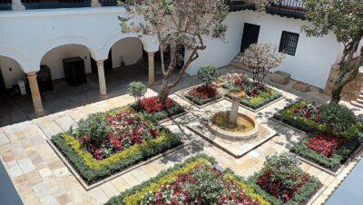 Patio central colonial del Museo Casa de Moneda en La Candelaria, Bogotá, con jardines florecidos, una fuente de piedra y corredores con arcos blancos