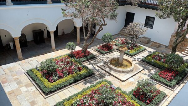 Patio central colonial del Museo Casa de Moneda en La Candelaria, Bogotá, con jardines florecidos, una fuente de piedra y corredores con arcos blancos